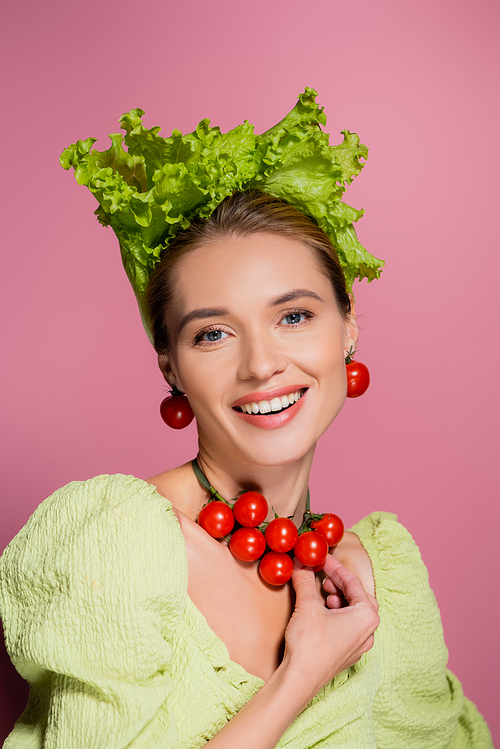 happy young woman in lettuce hat touching necklace made of cherry tomatoes on pink