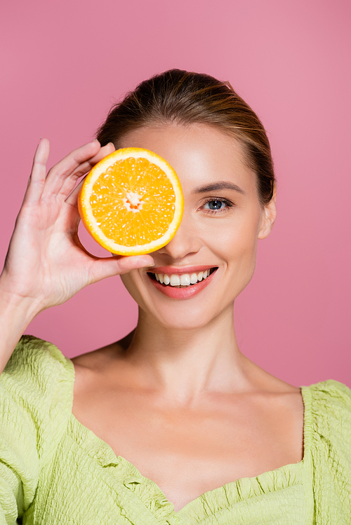 smiling woman covering eye with half of juicy orange isolated on pink