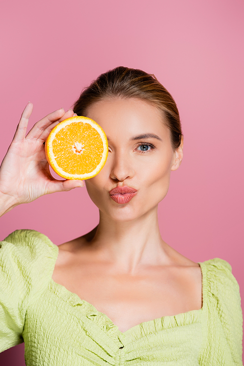 pretty woman sending air kiss while covering eye with orange half on pink