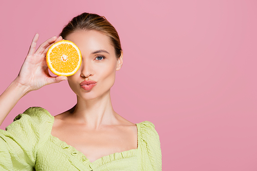 sensual woman covering eye with half of juicy orange and sending air kiss isolated on pink