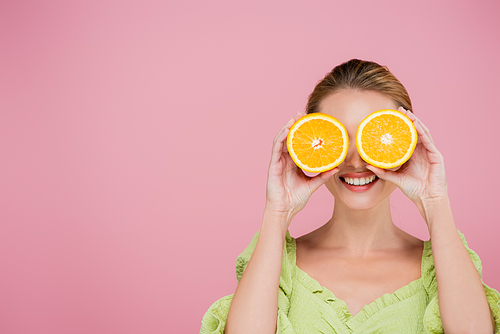 smiling woman covering eyes with halves of ripe orange isolated on pink