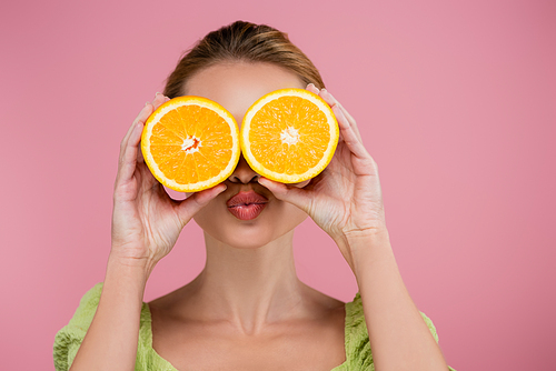 young woman sending air kiss while covering eyes with orange halves isolated on pink