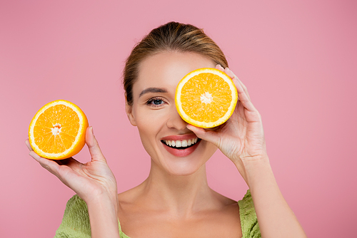 cheerful woman covering eye with half of ripe orange isolated on pink