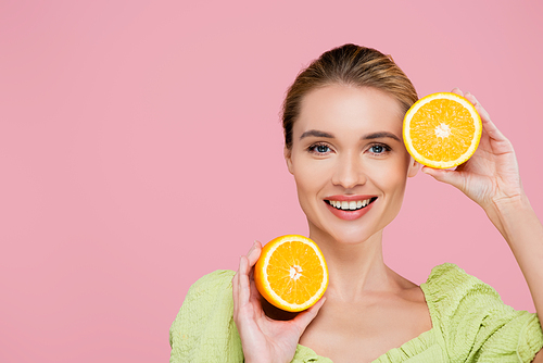 happy young woman smiling at camera while holding halves of orange isolated on pink
