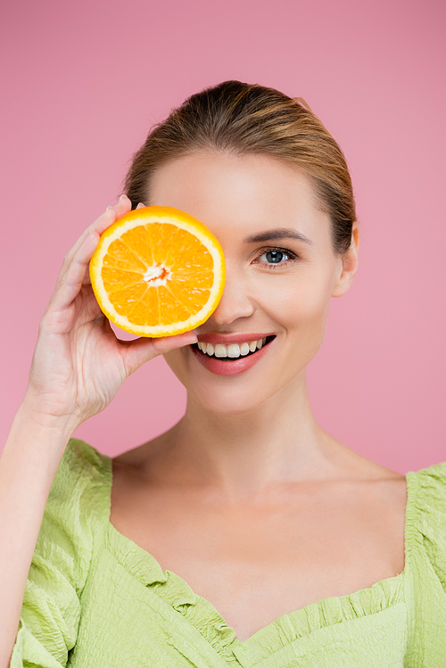 cheerful woman covering eye with half of juicy orange isolated on pink
