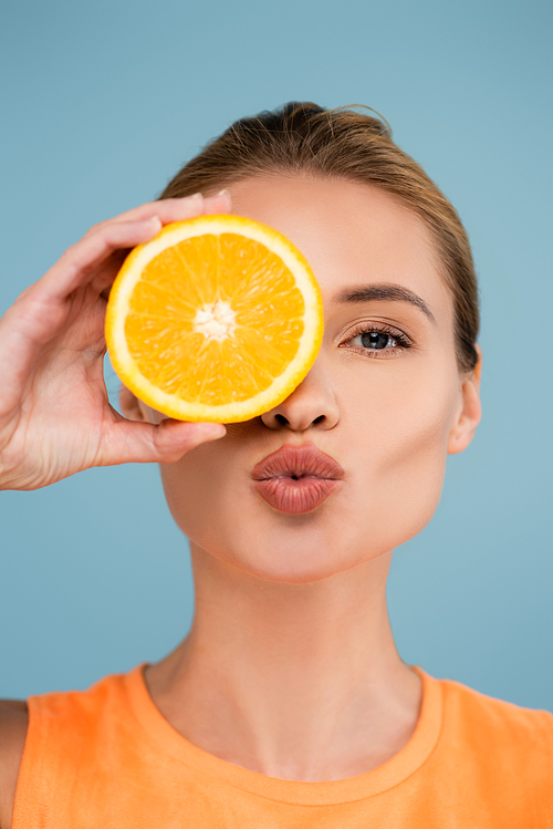 pretty woman covering eye with half of ripe orange while sending air kiss isolated on blue