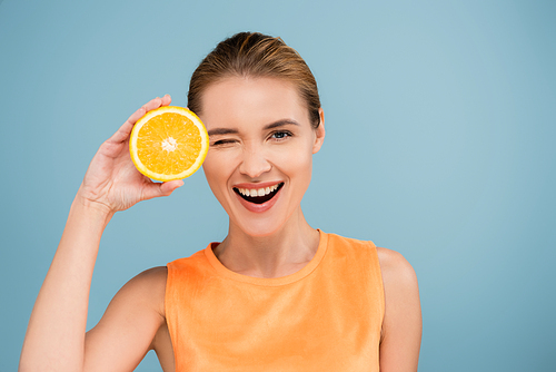 excited woman winking at camera while holding half of ripe orange isolated on blue