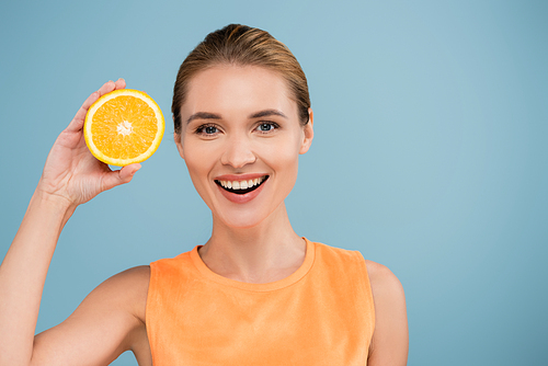 joyful woman smiling at camera while holding half of juicy orange isolated on blue