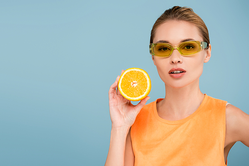 charming woman in colored eyeglasses posing with half of ripe orange isolated on blue