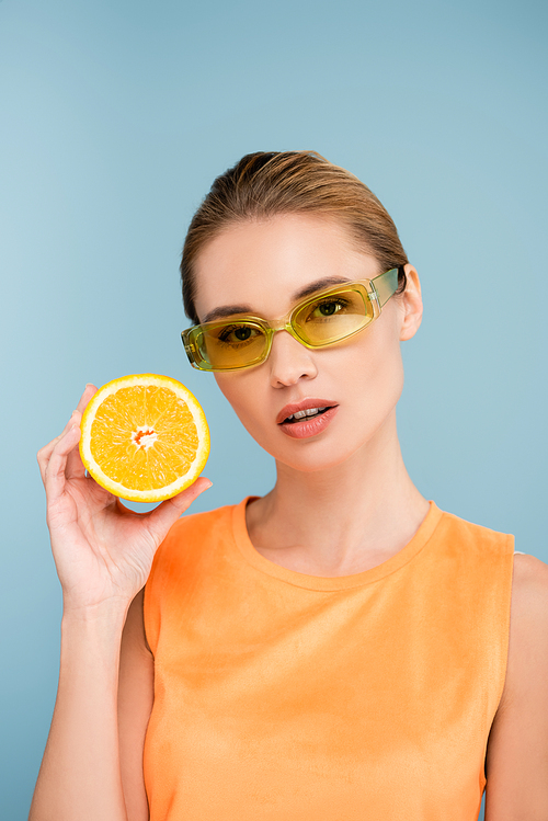 young woman in colored eyeglasses holding half of juicy orange isolated on blue