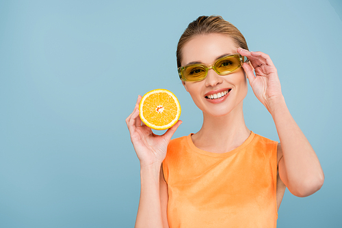 cheerful woman touching colorful eyeglasses while holding juicy orange isolated on blue