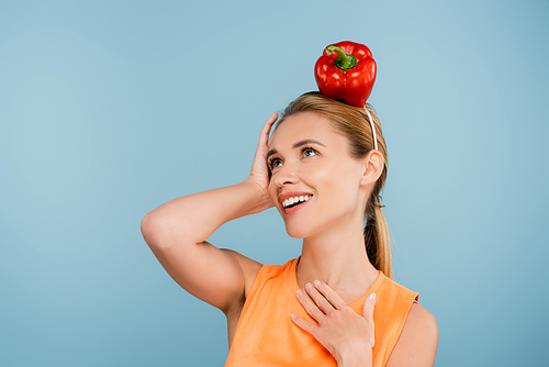 excited woman with red bell pepper instead of hat posing with hand on chest isolated on blue