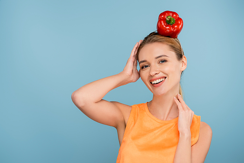 joyful woman with red bell pepper instead of hat smiling at camera isolated on blue