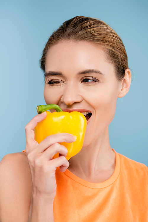 joyful woman biting ripe yellow bell pepper and winking isolated on blue