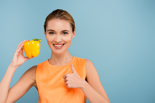 happy woman showing thumb up while holding yellow bell pepper isolated on blue