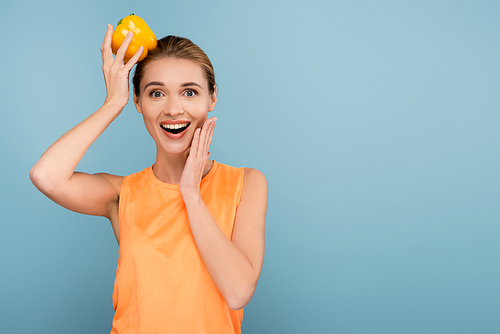 excited woman touching face while holding yellow bell pepper on head isolated on blue
