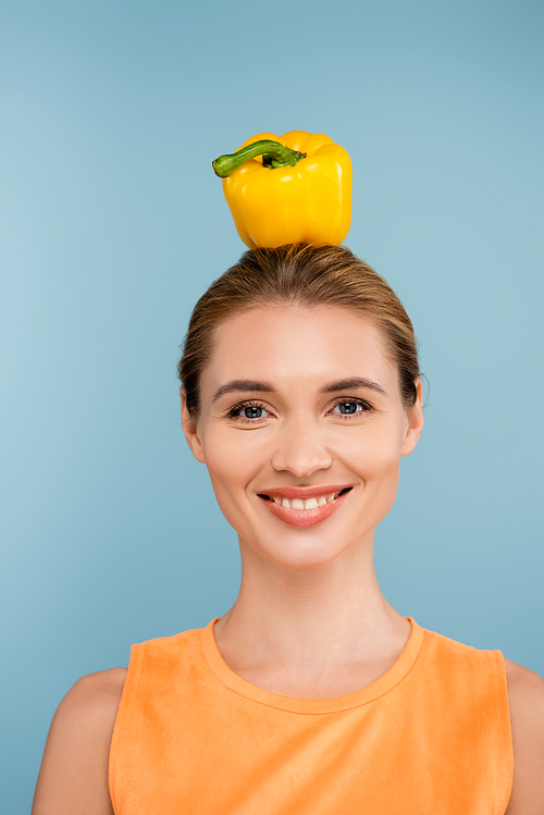 cheerful woman with yellow bell pepper on head smiling at camera isolated on blue