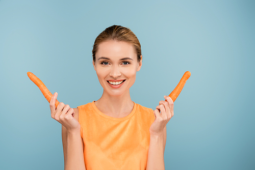 pretty woman smiling at camera while holding fresh carrots isolated on blue
