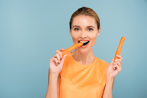 pleased woman  while eating whole carrot isolated on blue