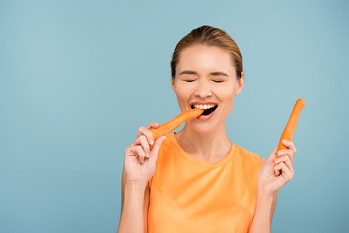 cheerful young woman with closed eyes biting whole carrot isolated on blue
