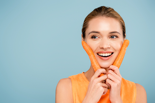happy woman with natural makeup holding fresh carrots near face isolated on blue