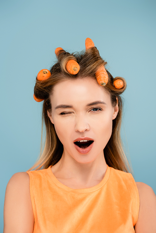 flirty woman with carrots instead of curlers winking at camera isolated on blue