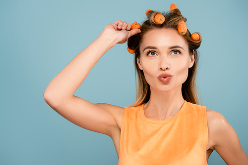 cheerful woman pouting lips while waving hair with fresh carrots isolated on blue