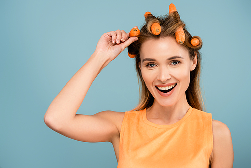 cheerful woman smiling at camera while curling hair with fresh carrots isolated on blue