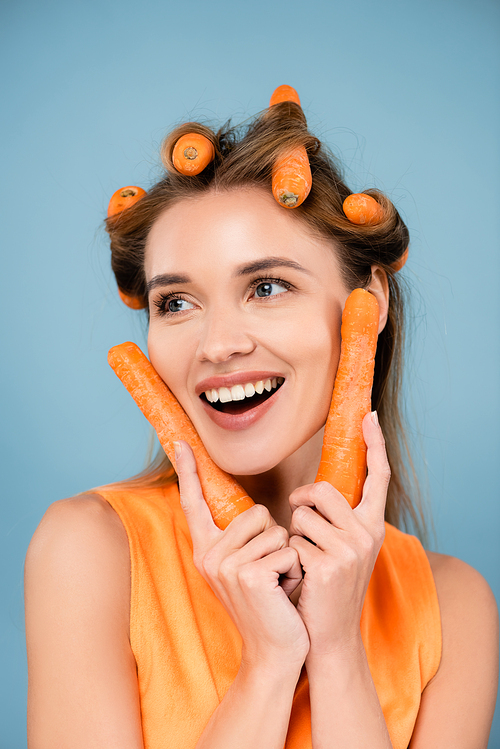 cheerful woman looking away while curling hair with fresh carrots isolated on blue