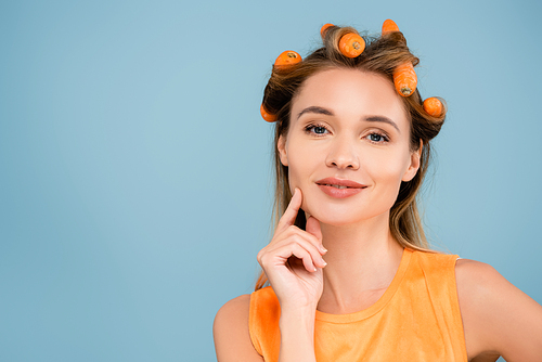 smiling woman with natural makeup touching face while waving hair with carrots isolated on blue
