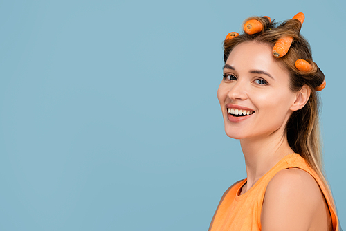 happy young woman using carrots instead of curlers isolated on blue