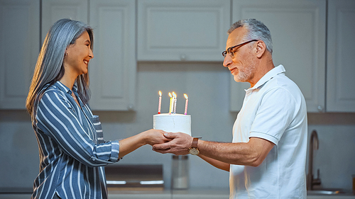 side view of pleased senior man taking birthday cake from happy asian wife