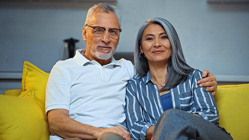 happy grey haired man embracing smiling asian wife while sitting on sofa at home