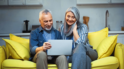 cheerful interracial couple sitting on sofa with laptop at home