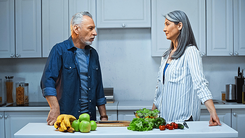 grey haired multicultural couple looking at each other while standing near fruits and vegetables in kitchen