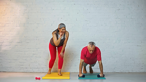 smiling asian woman pointing with finger near elderly husband doing push ups with dumbbells
