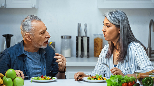 elderly multicultural couple talking during breakfast in kitchen