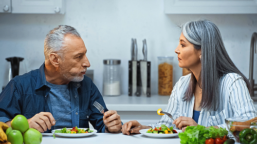 senior interracial couple talking while eating fresh salad in kitchen