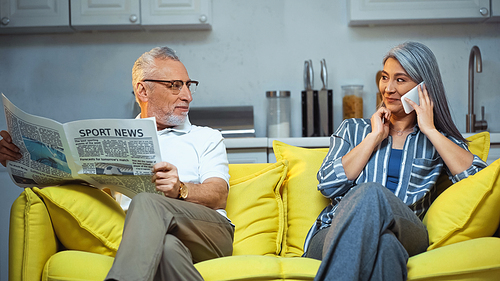 elderly man with newspaper looking at asian wife talking on smartphone