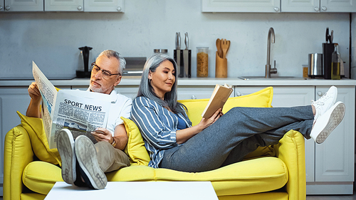 elderly interracial couple reading book and newspaper on sofa at home