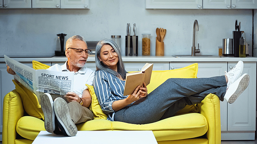 smiling asian woman reading book near senior husband with newspaper