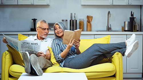 cheerful asian woman showing book to elderly husband holding newspaper