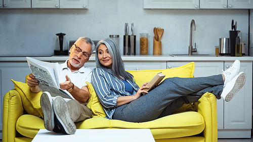 senior man pointing at newspaper near asian wife sitting on sofa with book
