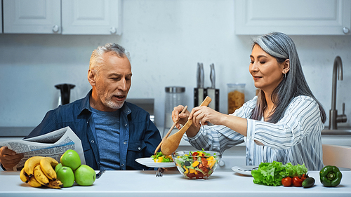 senior asian woman mixing salad near husband with newspaper in kitchen