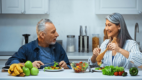 happy senior interracial couple talking during breakfast in kitchen