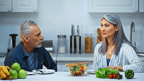 elderly interracial couple looking at each other during breakfast in kitchen