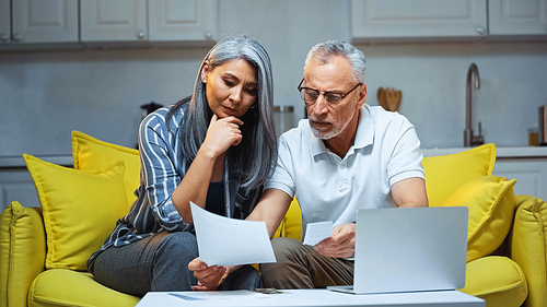 elderly man holding bills while sitting with asian wife near laptop