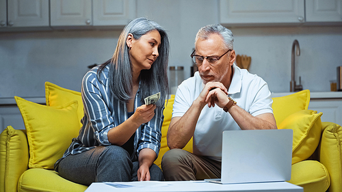 grey haired asian woman holding dollars near laptop and serious husband