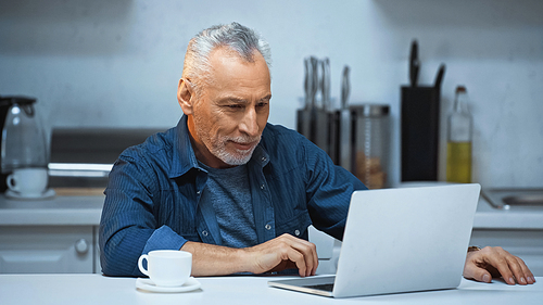 grey haired freelancer working near laptop and cup of coffee in kitchen