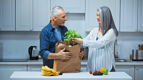 elderly interracial couple unpacking paper bag with fresh vegetables and fruits in kitchen